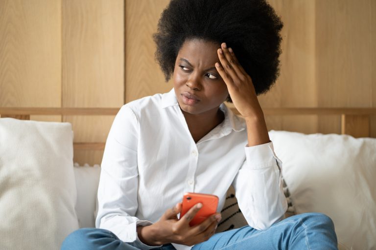 Sad Black woman sit on couch at home hold phone receive unpleasant breakup message, read bad news